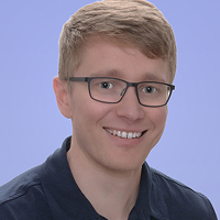 Smiling young man with short light brown hair and glasses wearing a dark shirt against a light purple background.