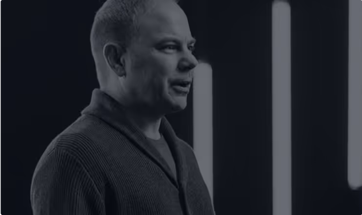 Man with short hair wearing a dark cardigan speaking against a background with vertical glowing light bars.