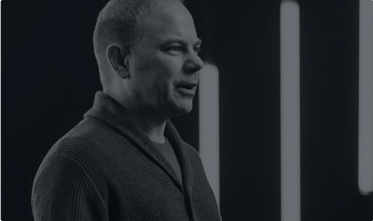 Man with short hair wearing a dark cardigan speaking against a background with vertical glowing light bars.
