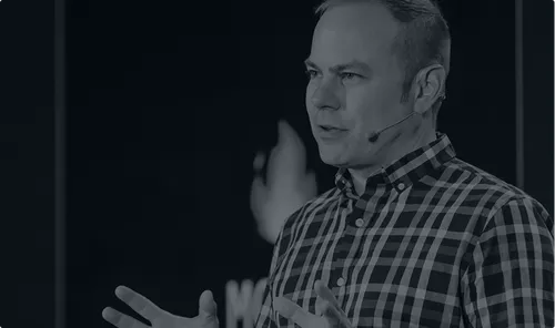 Man wearing a plaid shirt speaking with a headset microphone against a dark background.
