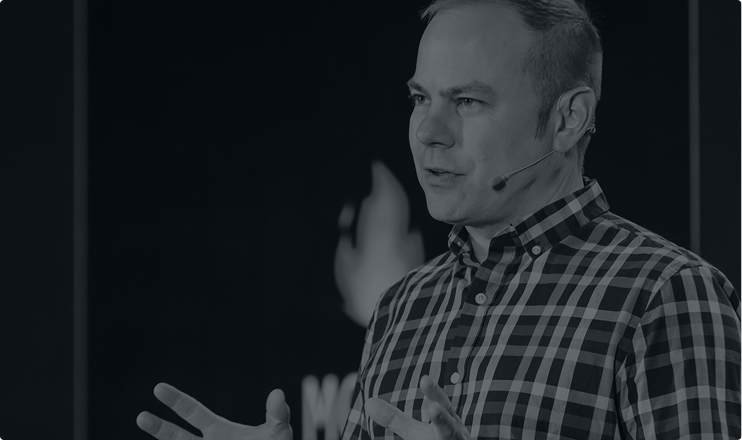 Man wearing a plaid shirt speaking with a headset microphone against a dark background.