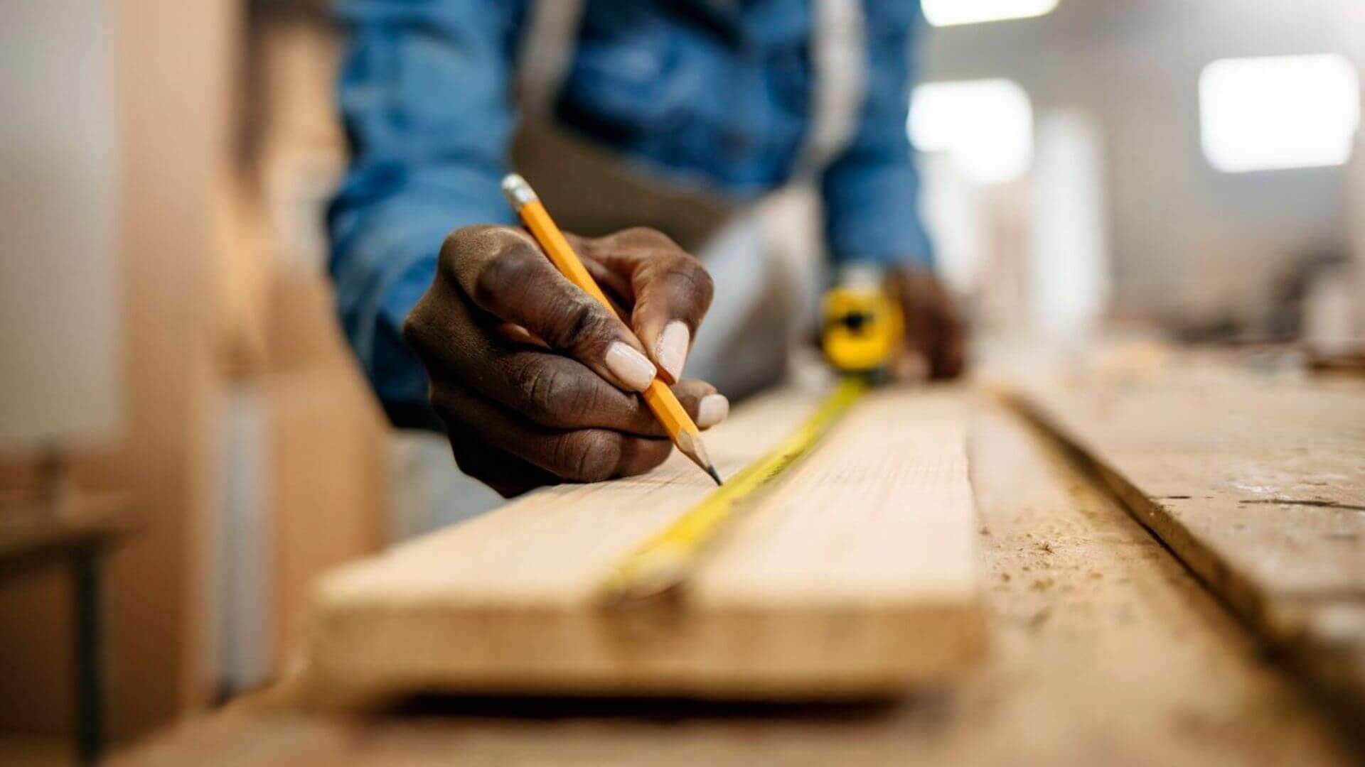 Person measuring a wooden plank with a yellow tape measure and marking it with a pencil.