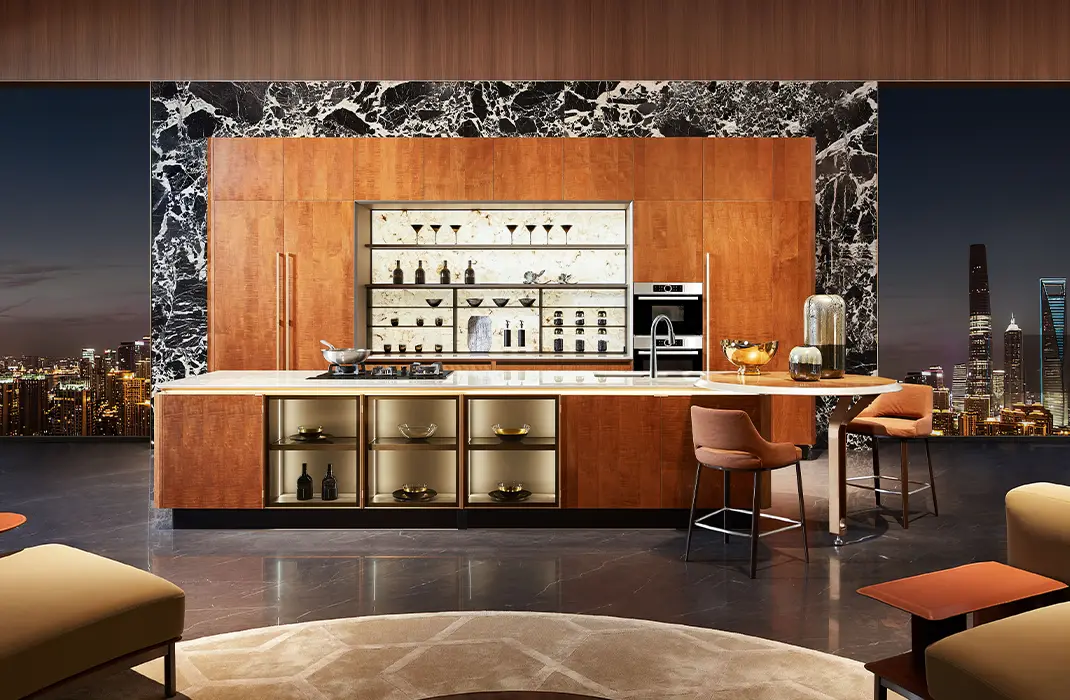 Contemporary luxury kitchen with cherry wood cabinetry, marble accents, large island with cooktop, and city skyline visible through floor-to-ceiling windows at night.
