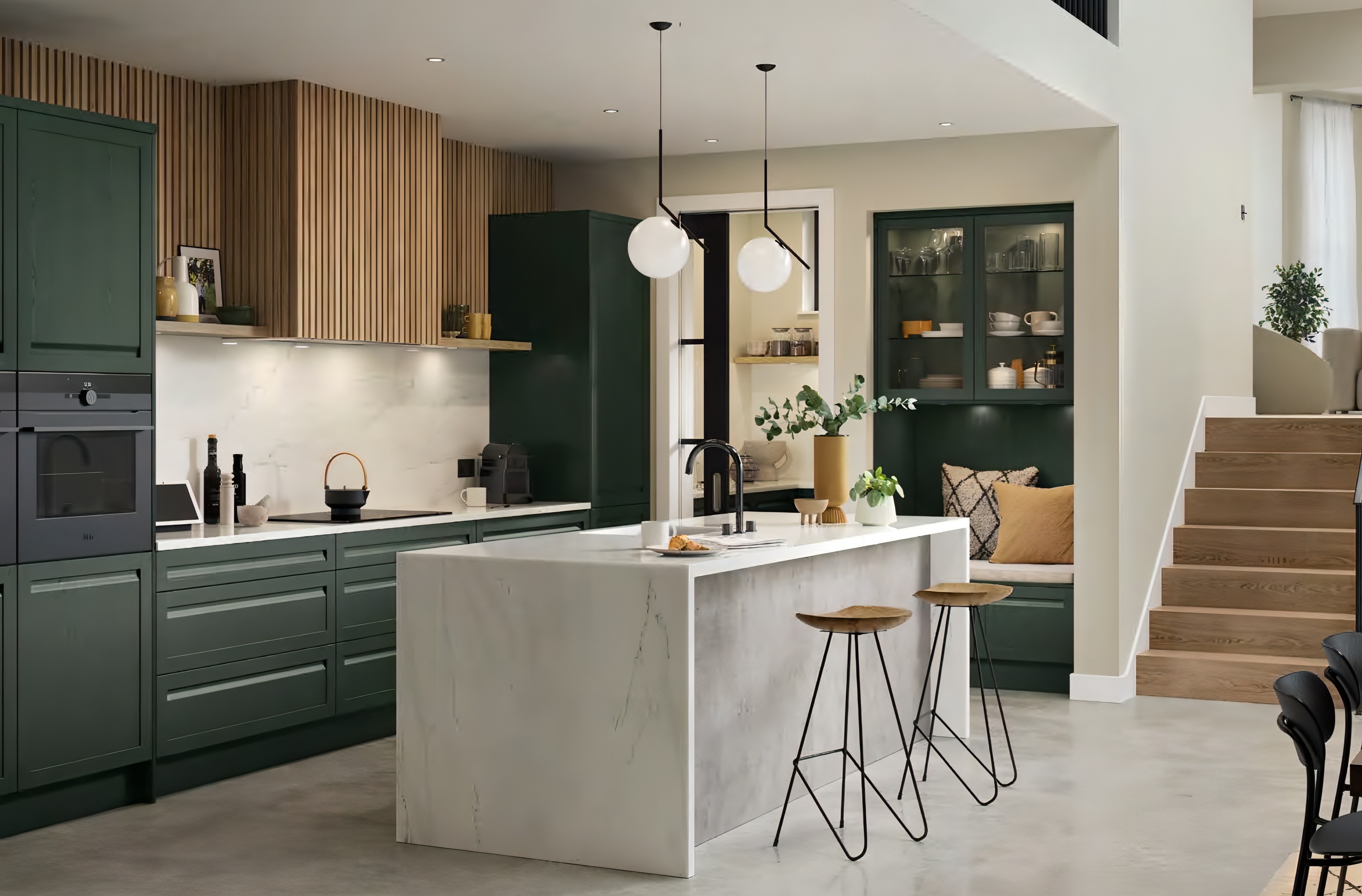 Modern kitchen with dark green cabinetry, a marble island with two wooden stools, pendant lights, and a cozy nook with pillows near wooden stairs.