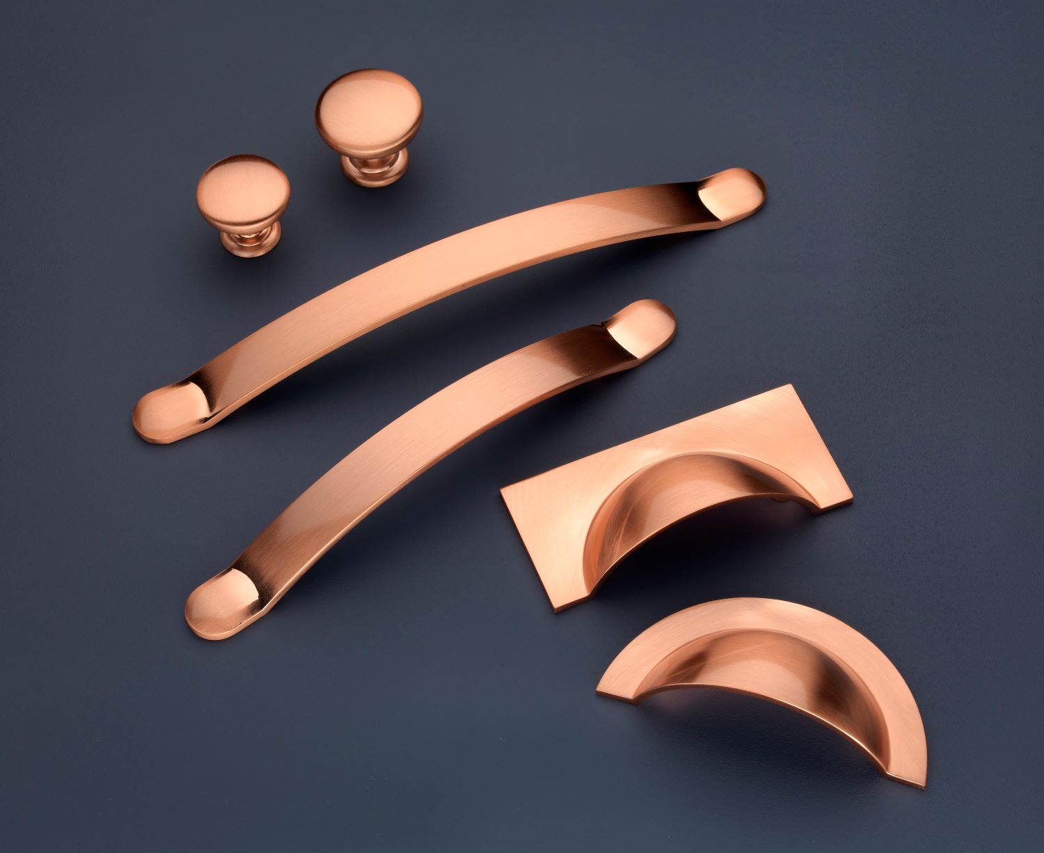 Copper-colored modern cabinet hardware including two round knobs, two curved handles, and two cup pull handles on a dark surface.
