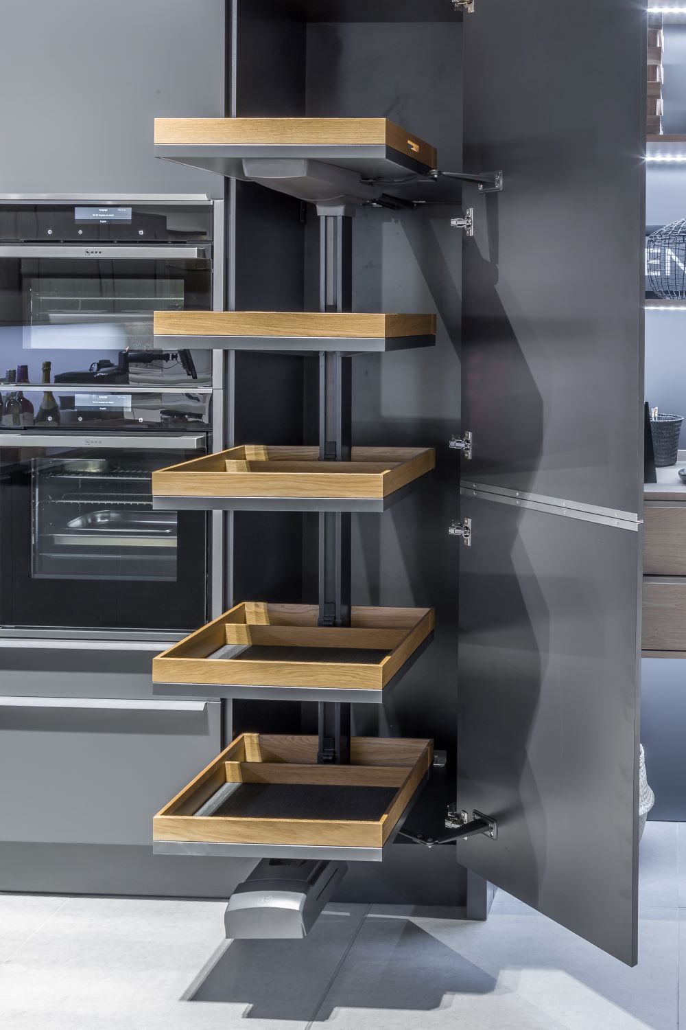 Modern kitchen cabinet with five pull-out wooden shelves and grey interior, next to built-in ovens.