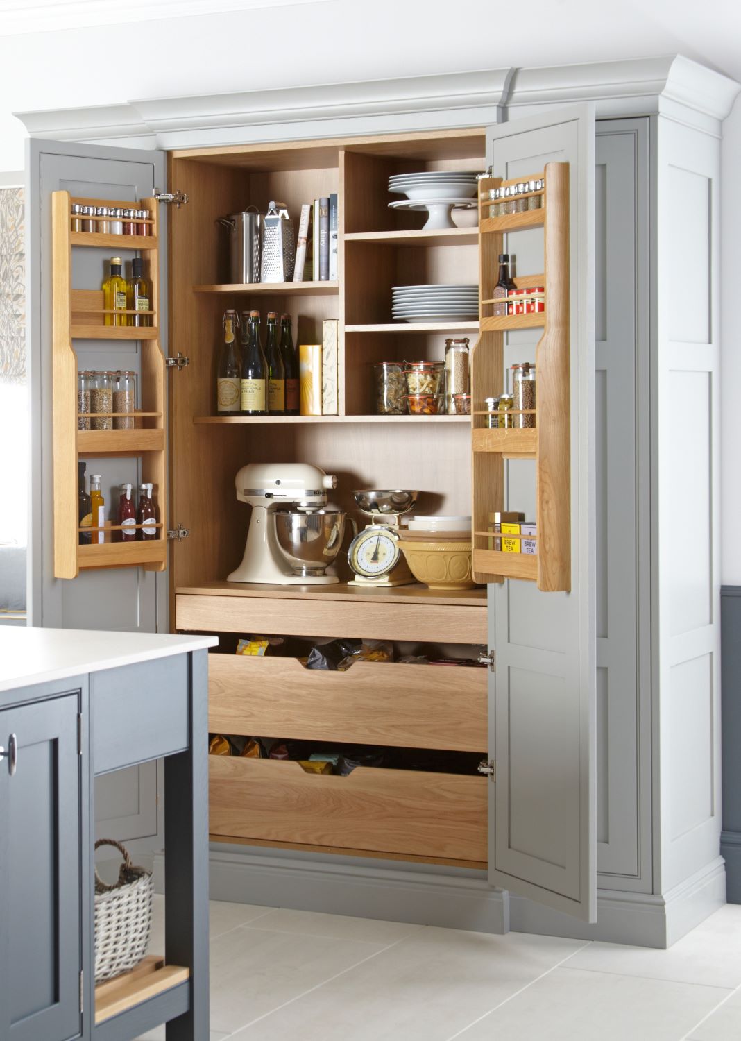 Open grey kitchen pantry cabinet with wooden interior shelves holding jars, bottles, plates, a stand mixer, kitchen scale, and storage drawers.