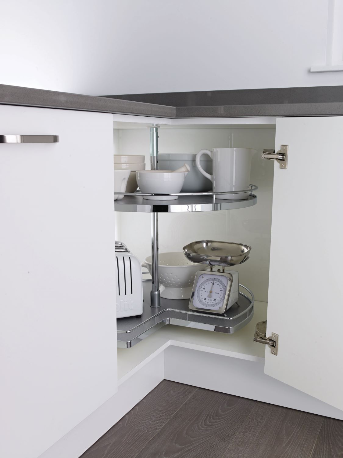 Corner kitchen cabinet with a two-tier lazy Susan holding white bowls, a white pitcher, a white toaster, a colander, and a kitchen scale.