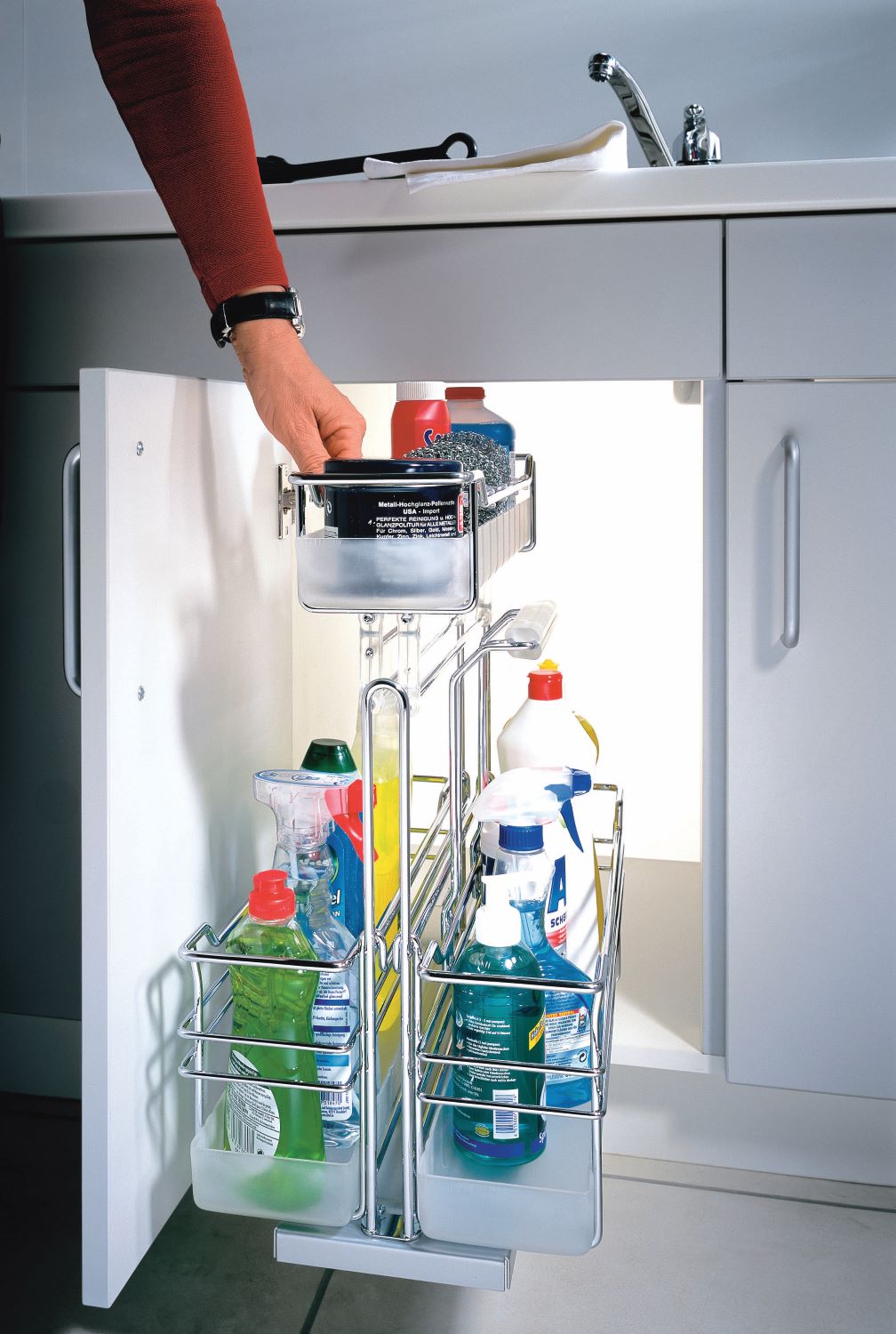 Pull-out metal organizer inside a white kitchen cabinet door holding various cleaning bottles and sponges, with a hand reaching in to grab an item.