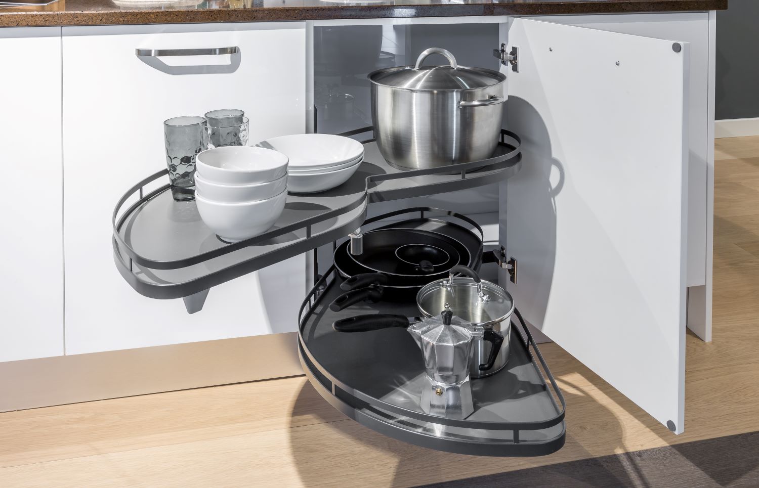 Corner kitchen cabinet with pull-out shelves holding pots, pans, glass cups, white bowls, and plates.