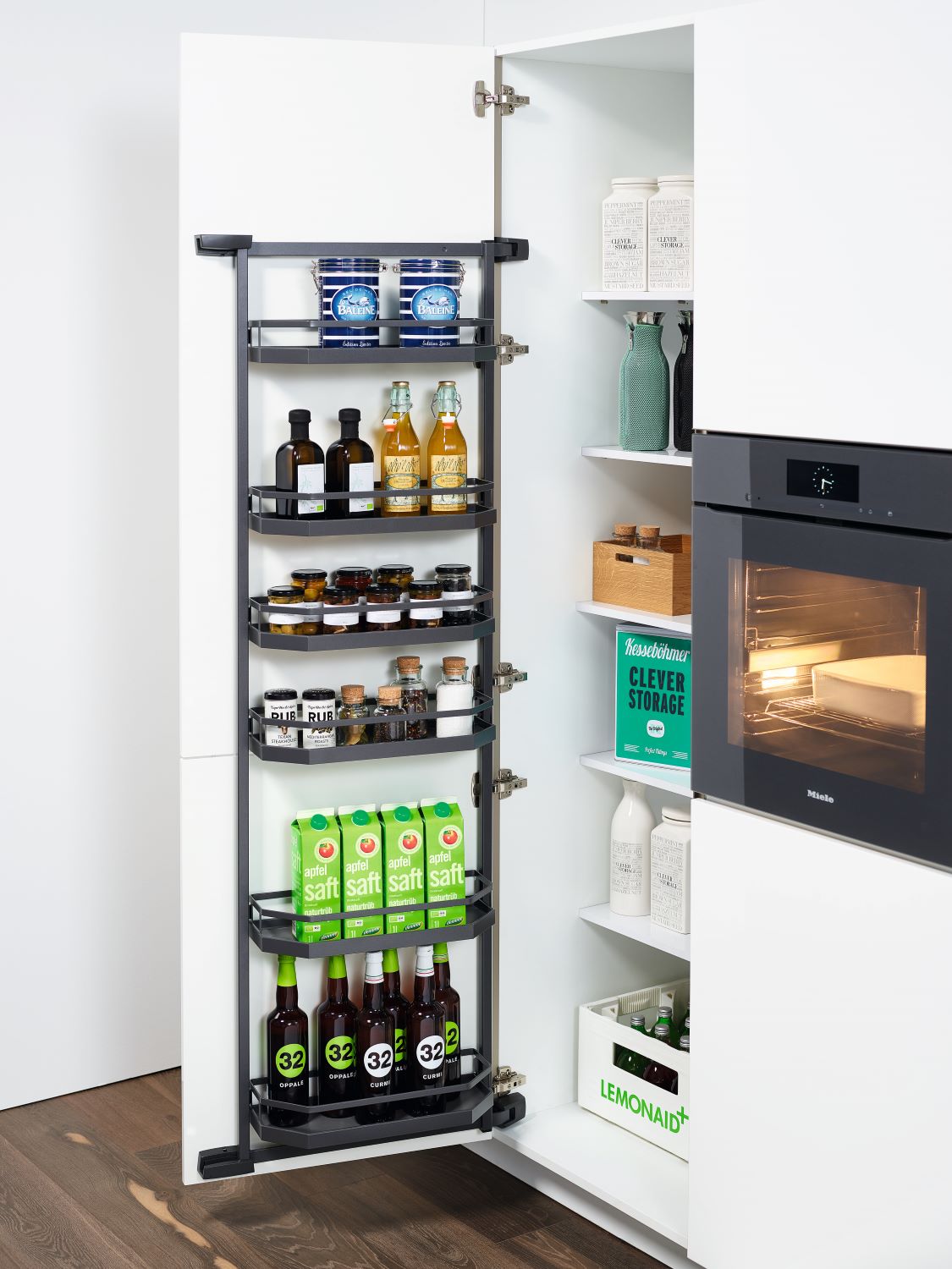 Open white pantry cabinet door with multiple black shelves holding jars, bottles, and cartons of apple juice, next to a built-in oven.