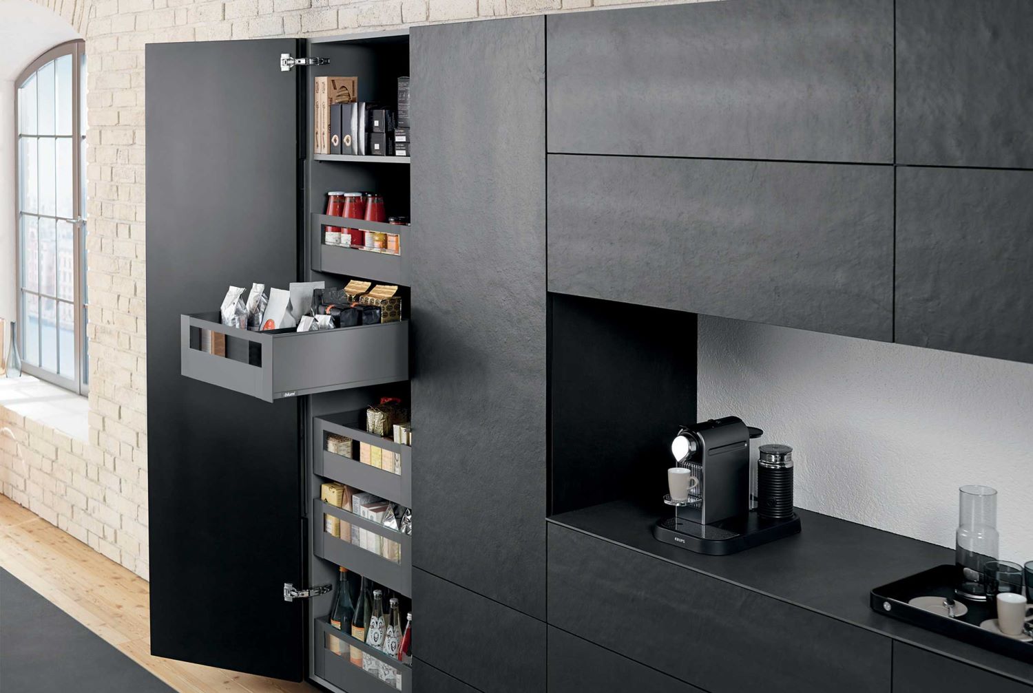 Modern kitchen with matte black cabinets, one cabinet door open revealing organized pull-out shelves stocked with jars, boxes, and bottles, next to a black espresso machine on the counter.
