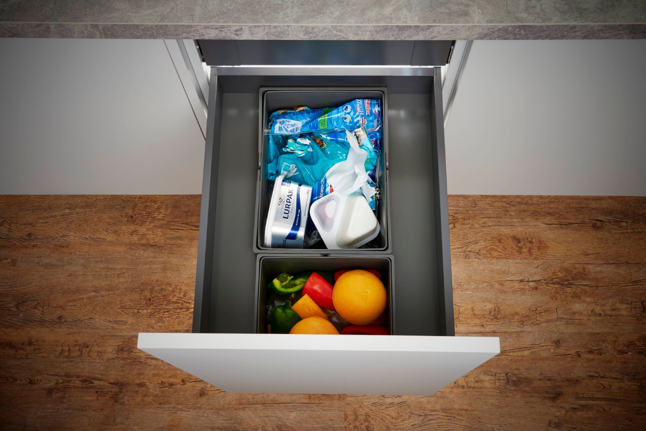 Open kitchen drawer with two compartments, one filled with packaged dairy products and the other with fresh vegetables and oranges.