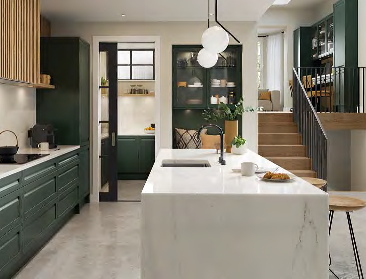 Modern kitchen with dark green cabinets, white marble island with sink, hanging globe lights, and wooden stools, next to a stairway leading to a sitting area.
