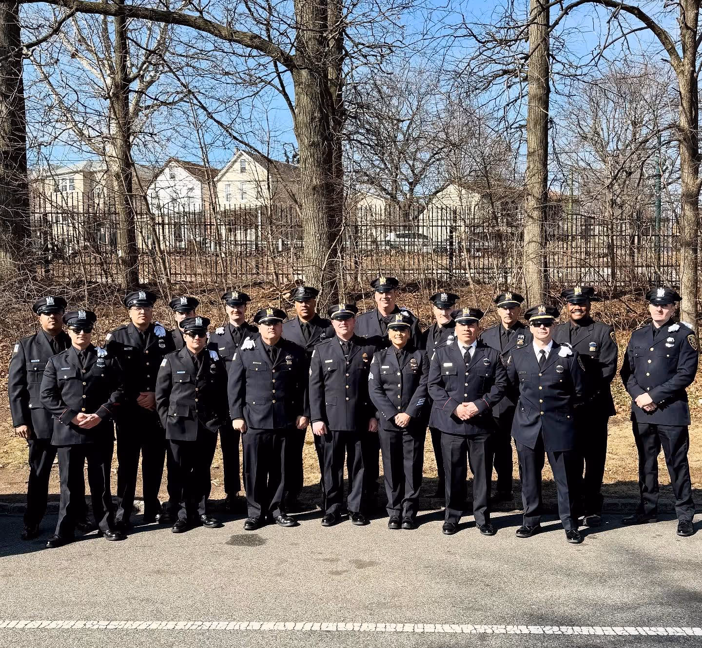 Group of uniformed police officers posing together outdoors in formal dress uniforms. They stand in rows on a paved surface with leafless trees, houses, and a black metal fence in the background. The officers wear dark navy uniforms, caps, and badges, signifying participation in a law enforcement memorial or ceremonial event honoring fallen colleagues.