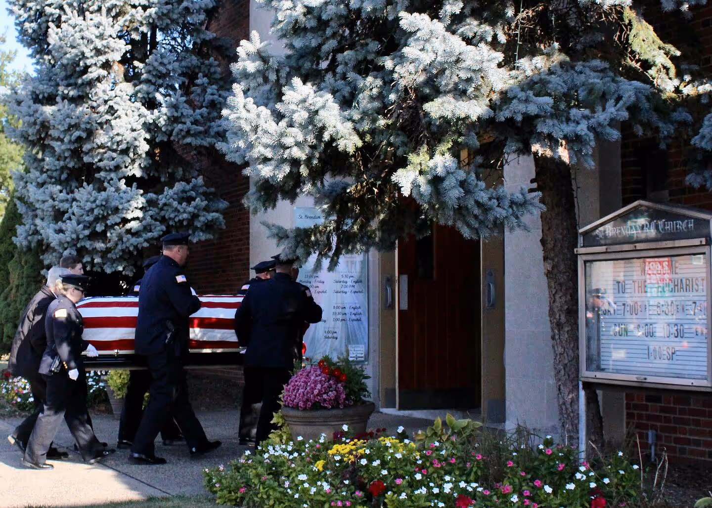 Uniformed police officers carry a flag-draped casket into St. Brendan Church for a funeral service, with colorful flowers arranged outside the entrance.