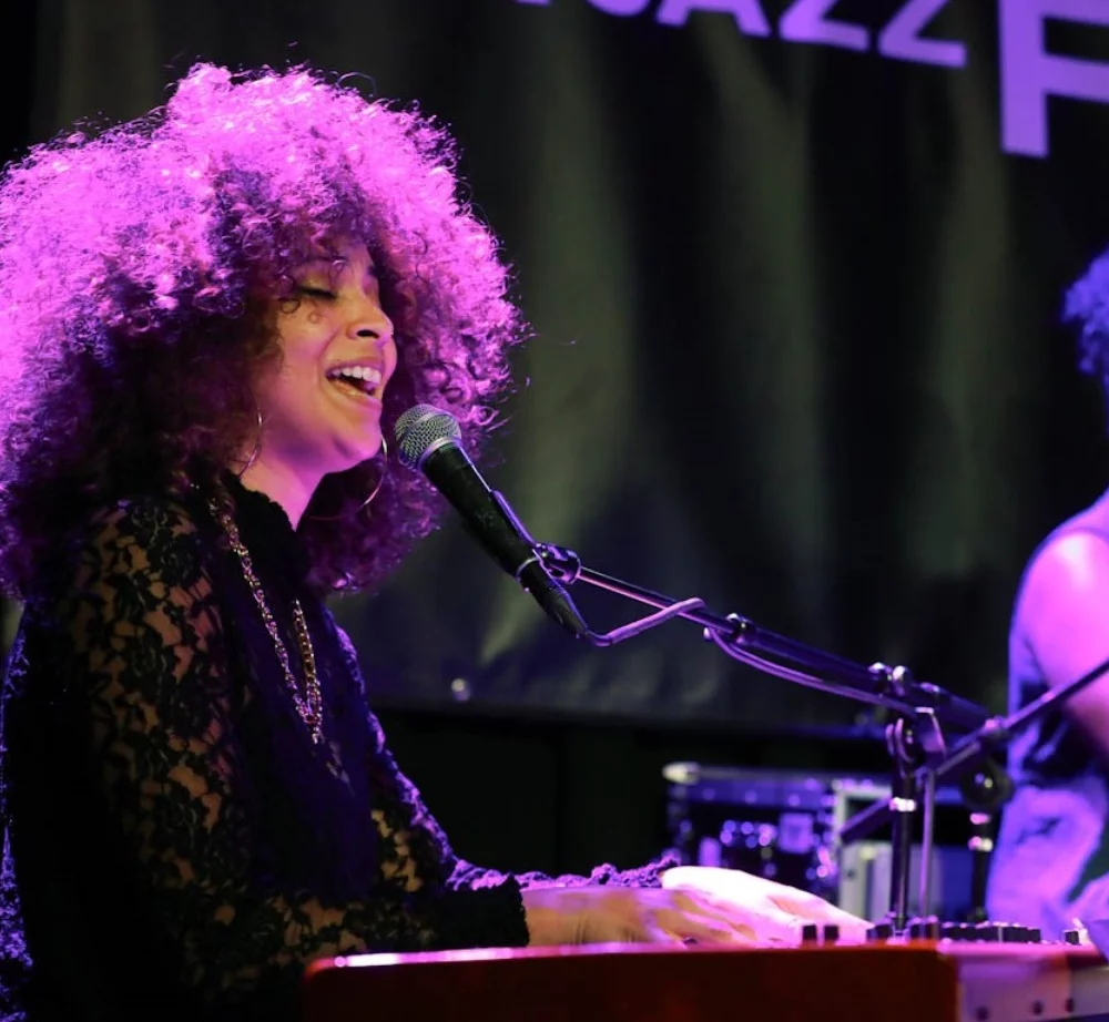 African American woman jazz singer Esperanza Spaulding backlit with purple lighting singing into a microphone while seated at the piano.