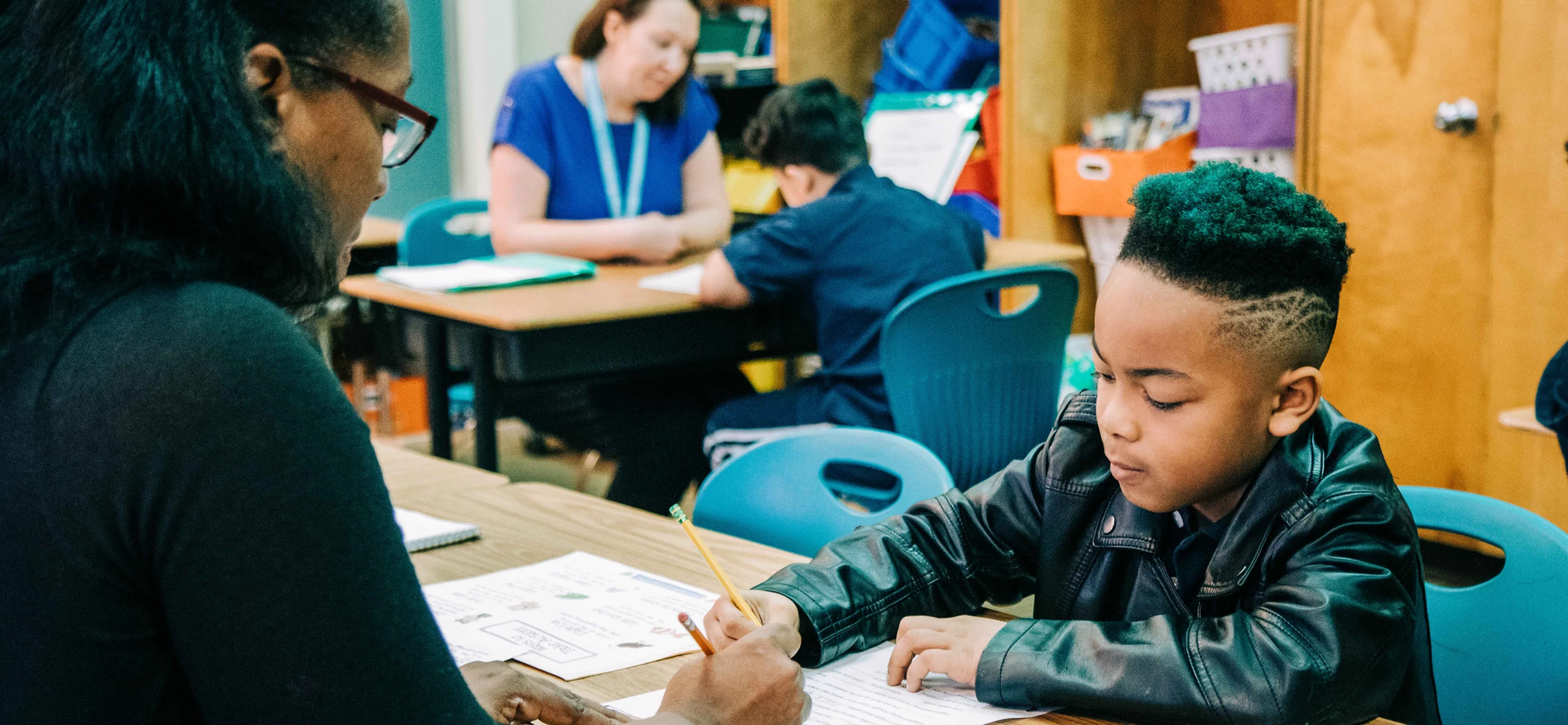 A student sitting at a table with a teacher, both are writing with a pencil and looking down at their paper. 