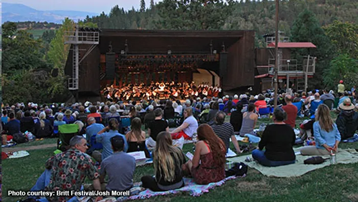 A large audience on blankets in front of an outdoor stage with an orchestra