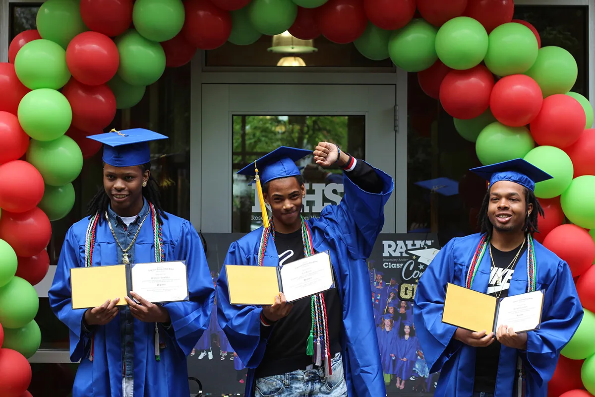 3 high school graduates in blue gowns and mortarboards holding their diplomas, surrounded by a green and red balloon arch
