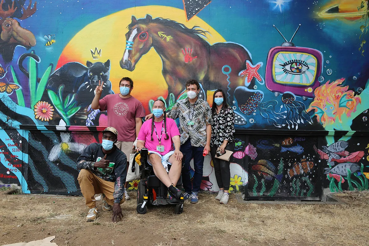 5 artists sitting and standing in front of a multi-colored wall mural; photo courtesy of Vibe of Portland