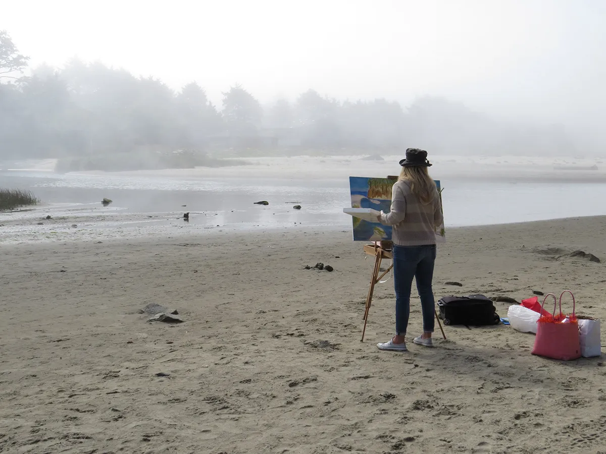 A woman painter standing on the beach in front of an unfinished canvas.