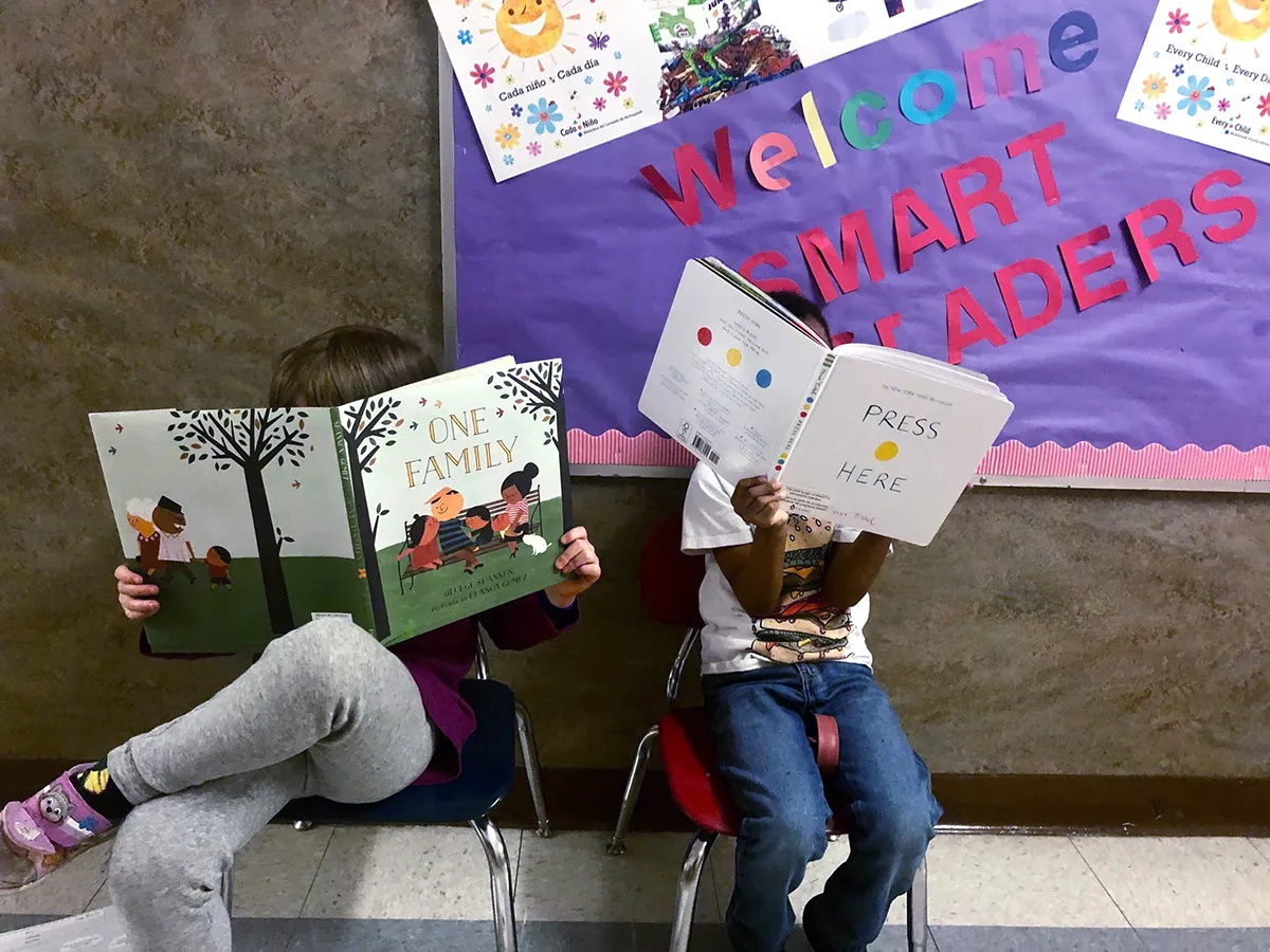 2 young students sitting on small chairs, they are reading and their faces are obscured by the books; Photo courtesy of SMART Reading