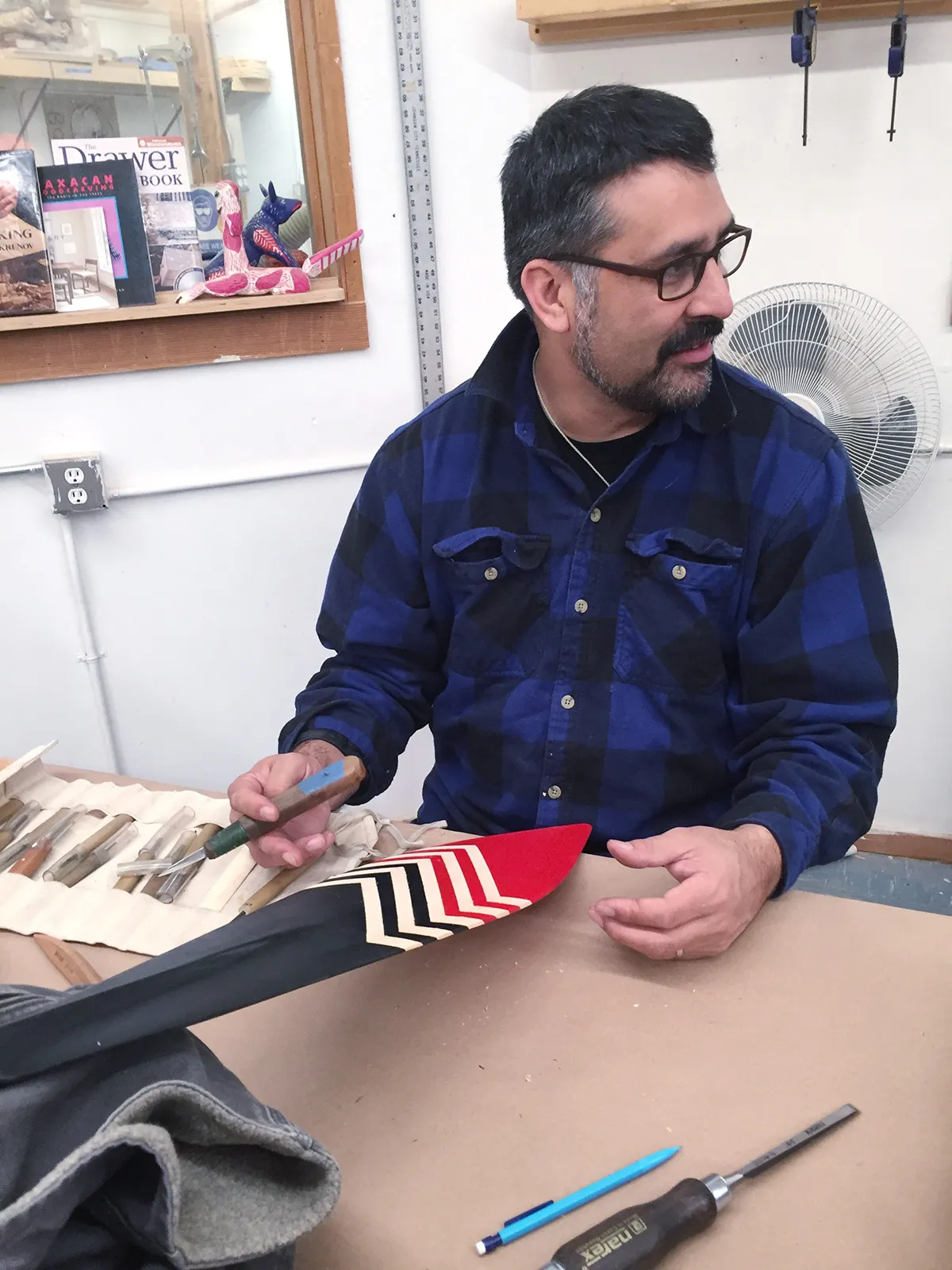  A carver in a blue plaid shirt, working on a paddle with a chevron pattern in black, cream and red; Photo courtesy of Native Arts and Cultures Foundation / Artist: Shirod Younker