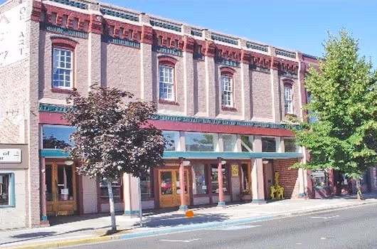 Exterior of the Historic G Street building houing Grants Pass Museum of Art, a two story brick building with storefronts opening onto a sidewalk with trees