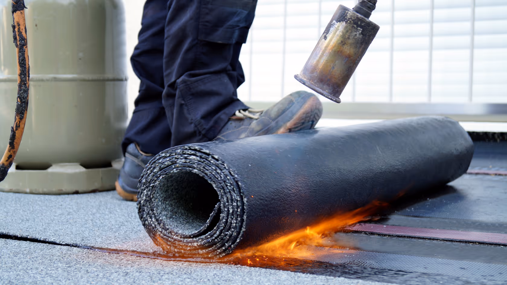 Worker applying torch-down roofing membrane with a flame torch on a rooftop surface.
