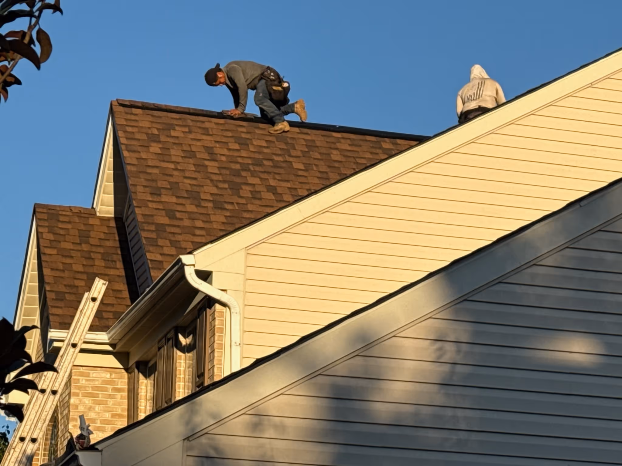 Two workers on a roof under clear blue sky, one crouching and the other sitting, with a ladder leaning against the house.