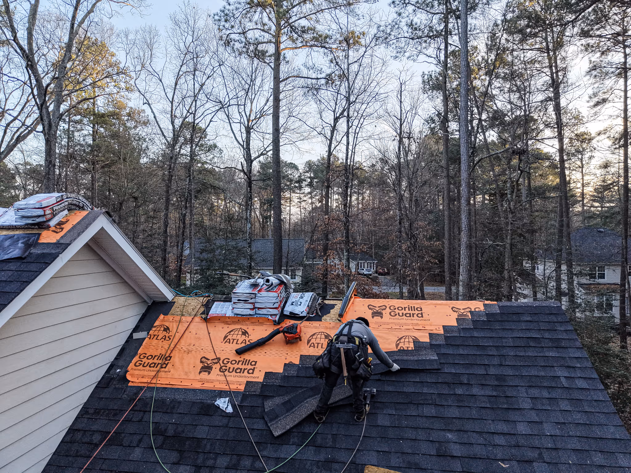 Roofer installing dark asphalt shingles on a house roof with orange Gorilla Guard underlayment, surrounded by trees.