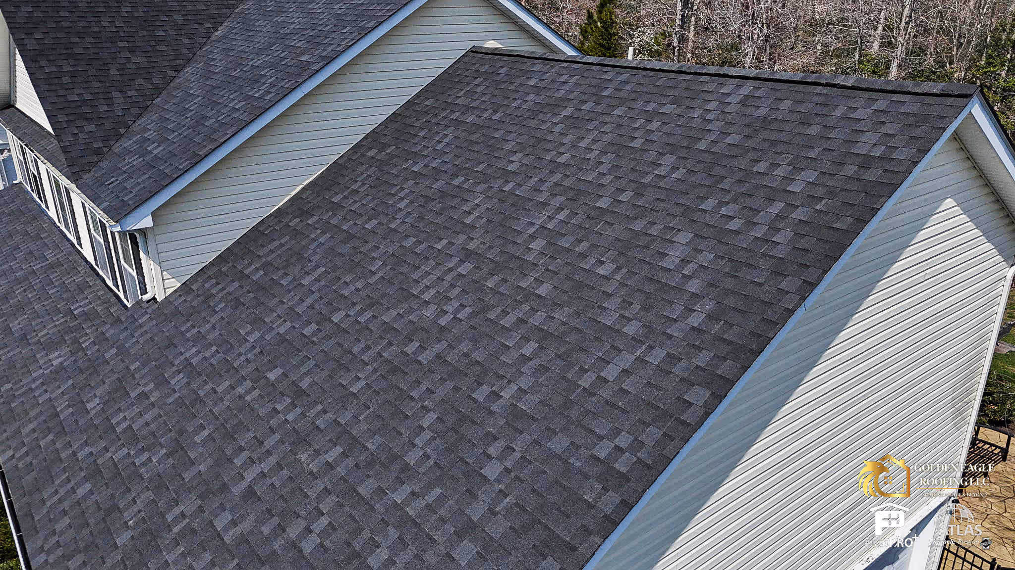 Dark gray shingle roof on a beige siding house viewed from above with bare trees in the background.