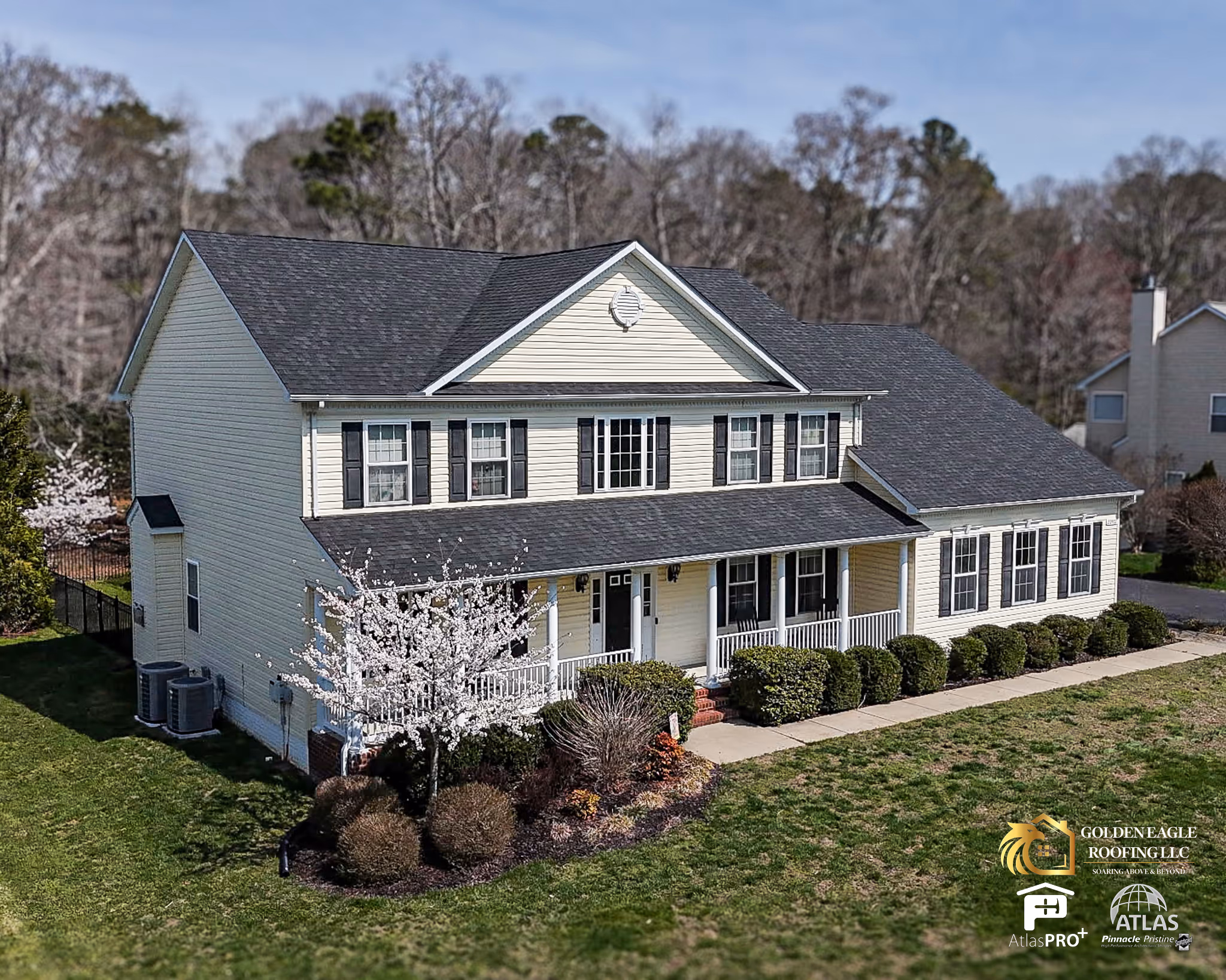 Two-story yellow house with black shutters, a dark gray roof, and a front porch surrounded by trimmed bushes and a blooming tree.
