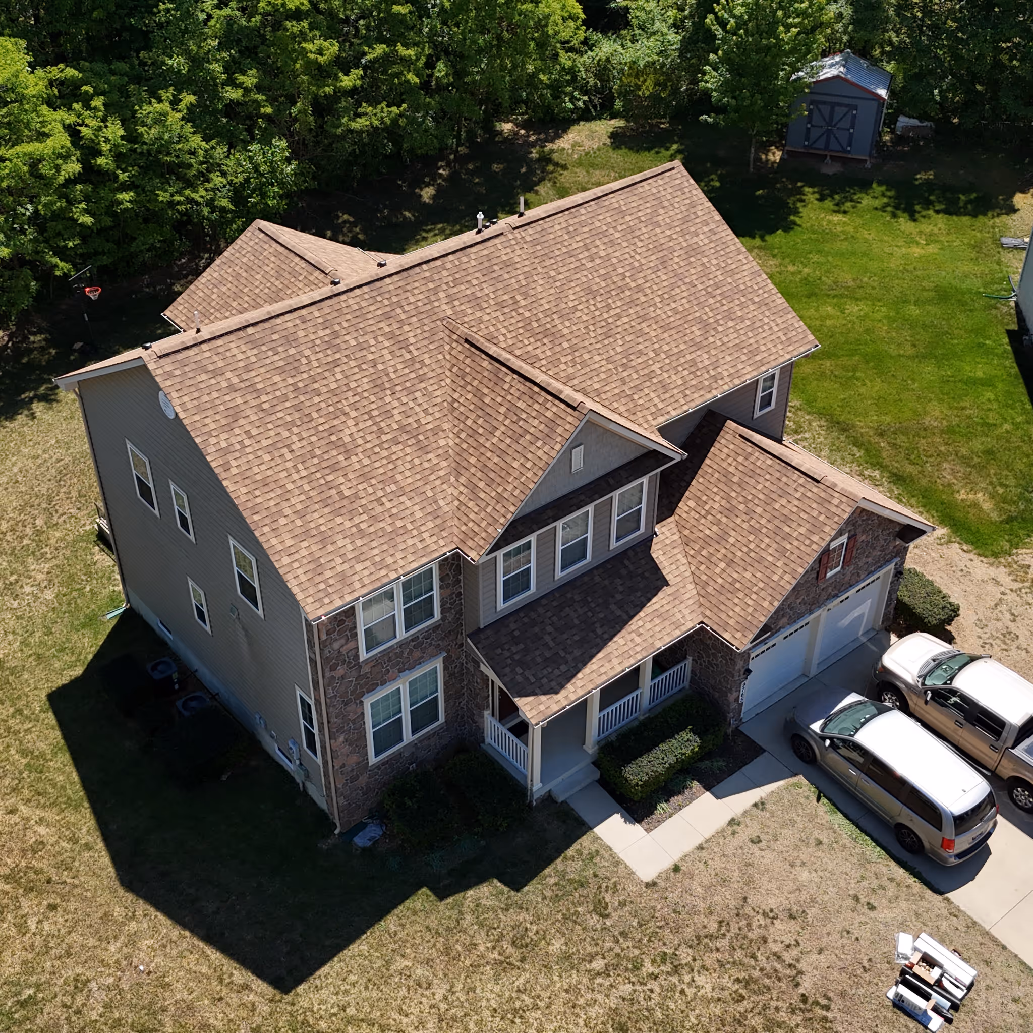 A two-story house with a brown shingle roof, stone facade, front porch, and two cars in the driveway.