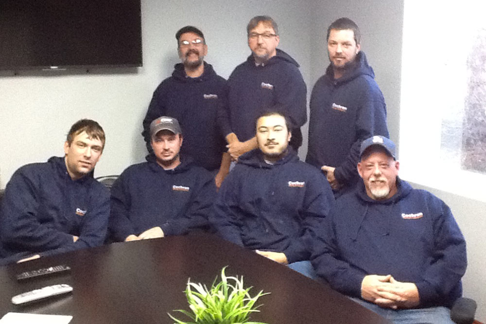 Group of seven men wearing navy hoodies with company logo, seated and standing around a conference table with a small green plant.