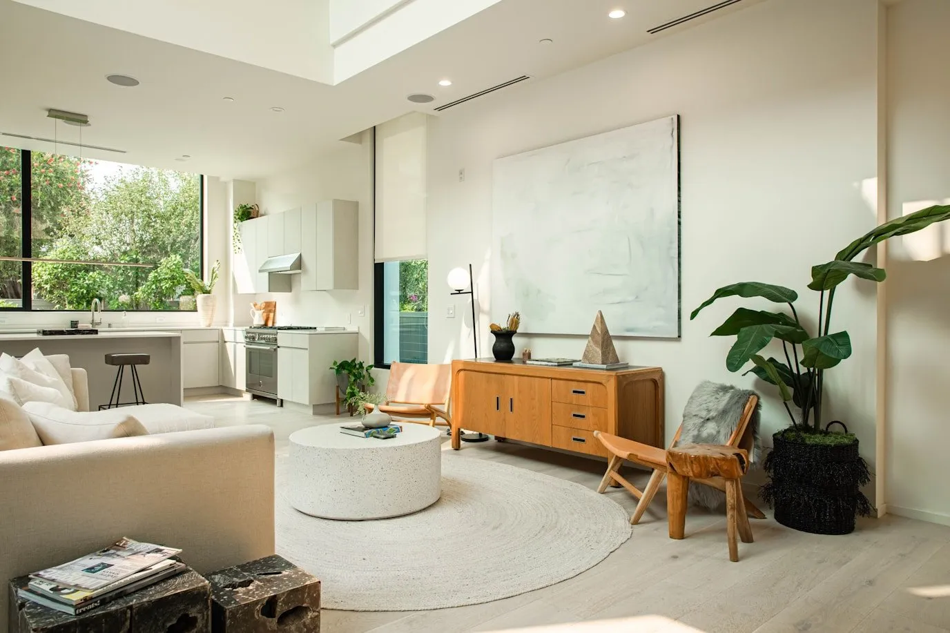 Bright modern living room with beige sofa, round white coffee table on a circular rug, wooden sideboard, leather chair, and large windows showing green trees outside.
