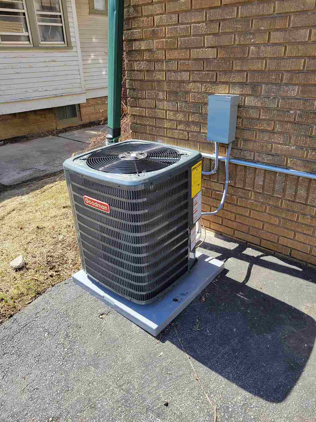 Goodman brand outdoor air conditioning unit installed on a concrete slab beside a brick house wall.