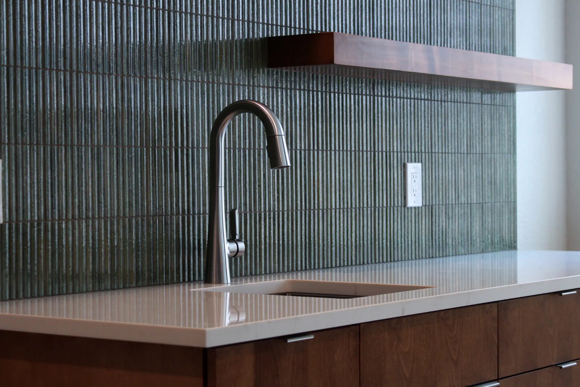 Close-up of wet bar sink and faucet against a glossy green ribbed tile backsplash, showing the smooth white countertop and dark wood drawers below.
