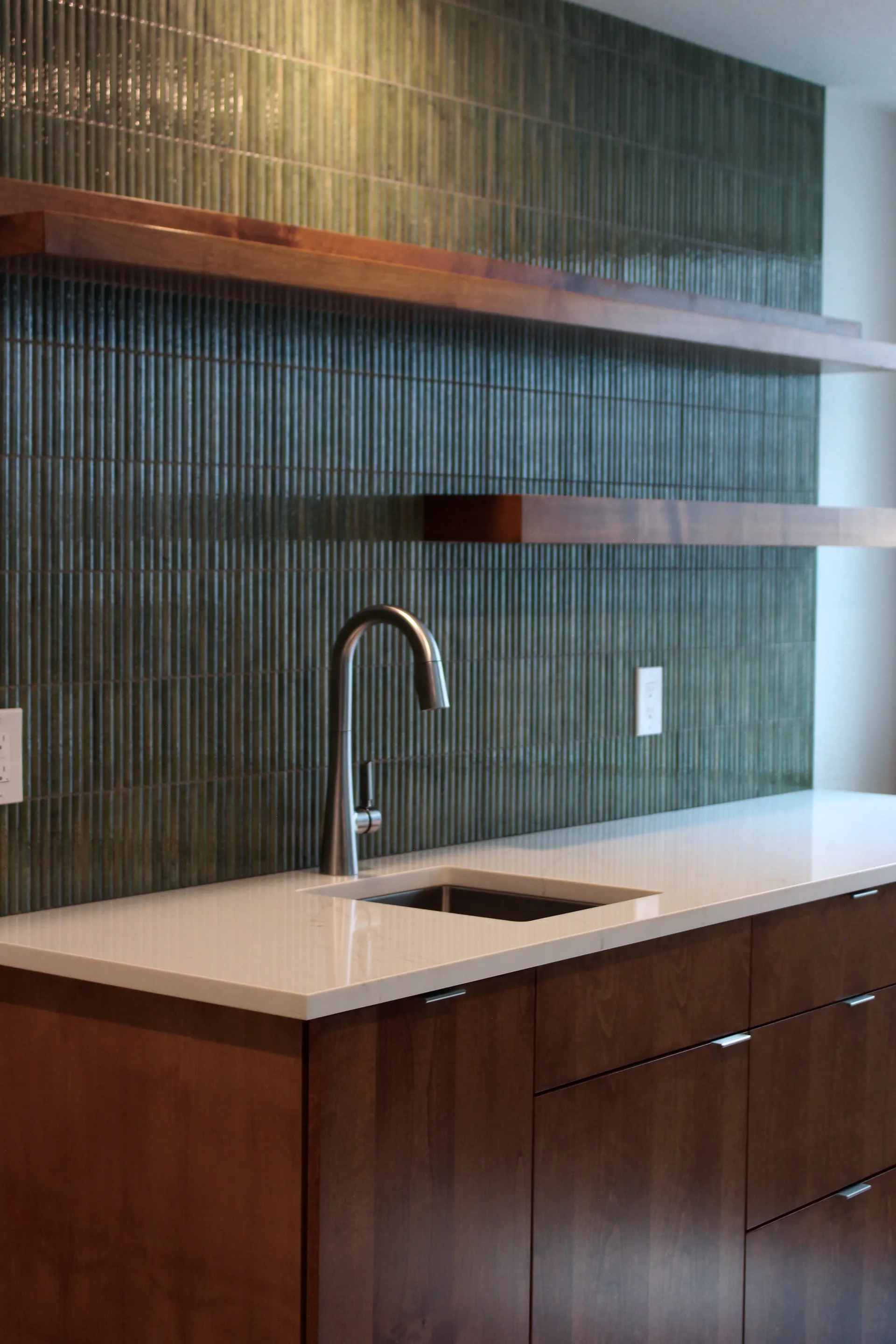 Perspective view of wet bar corner showing faucet, sink, and warm wood tones against the textured green backsplash.