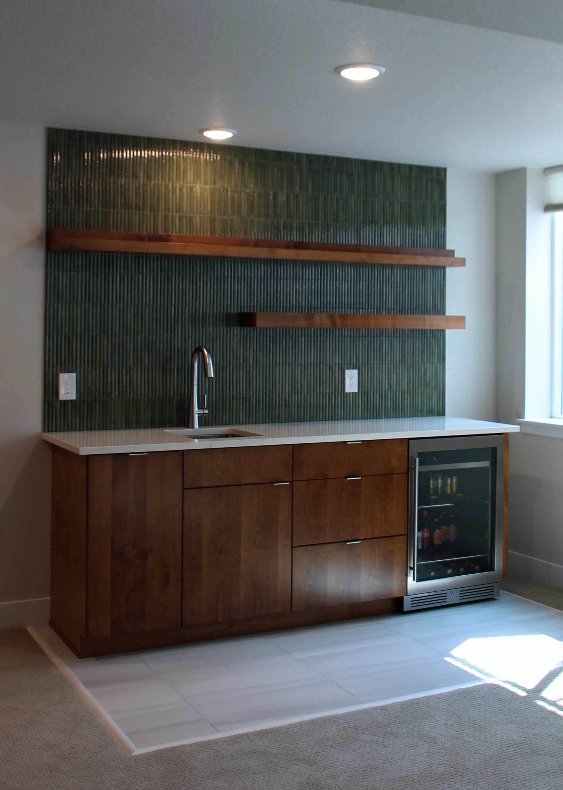 Angled view of wet bar showing floating wood shelves, green tile backsplash, and integrated beverage fridge.