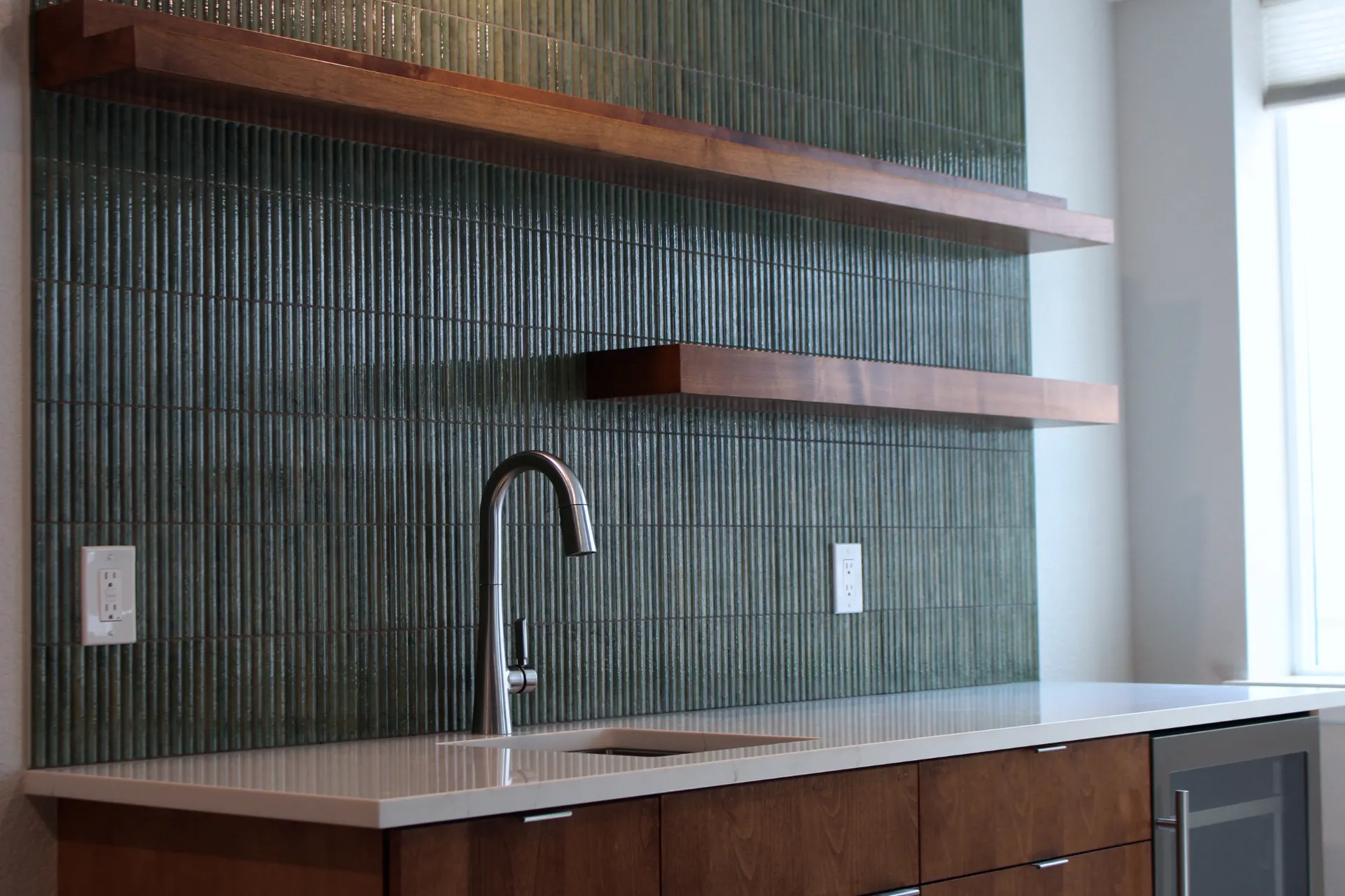 Perspective view of wet bar corner showing faucet, sink, and warm wood tones against the textured green backsplash.