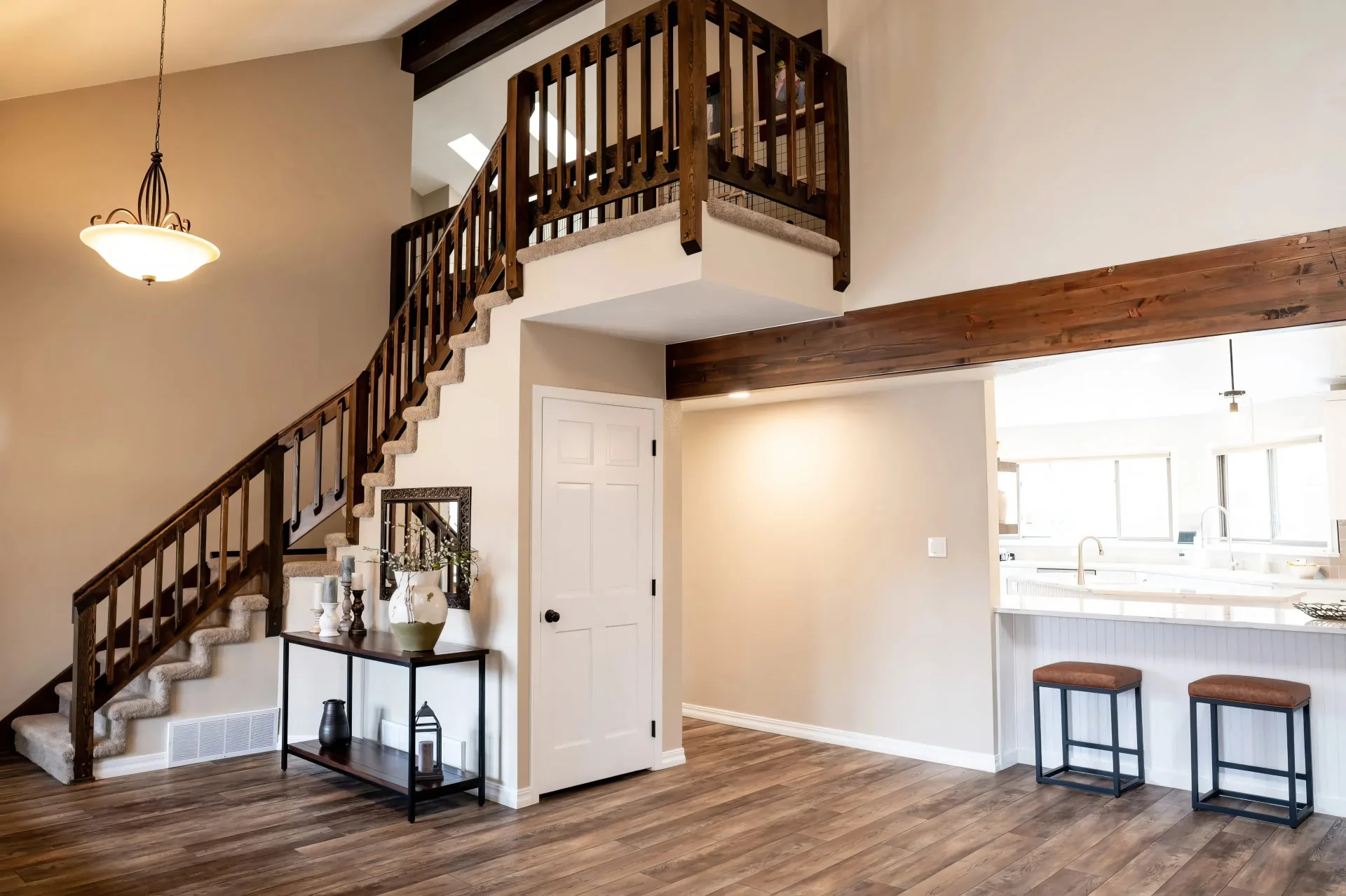 Spacious living area with wooden flooring and a staircase leading to an upper level. A modern light fixture hangs from the ceiling, and the open kitchen features stools by a counter. The decor is warm and inviting.
