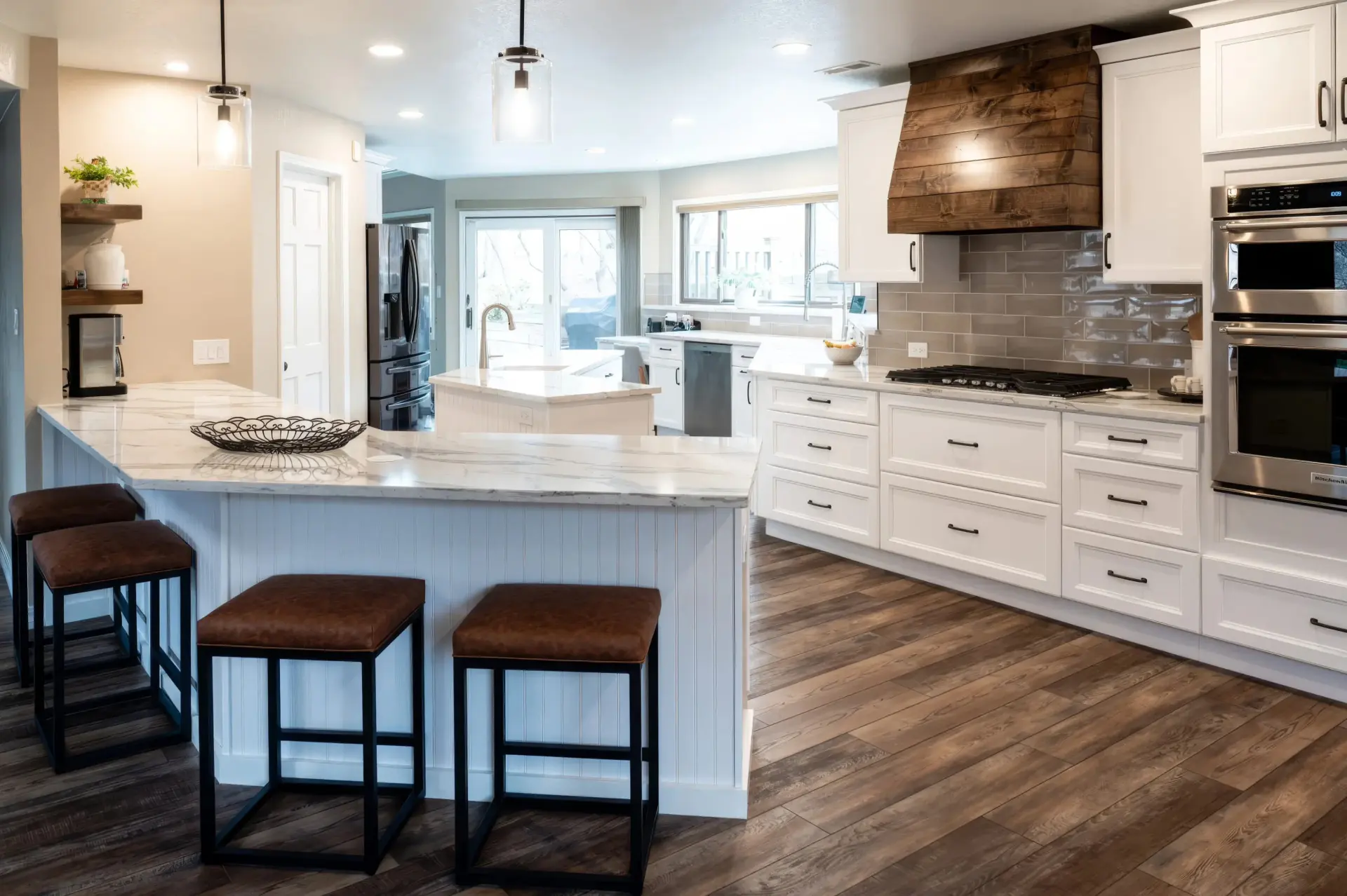 Modern kitchen with white cabinets, marble countertops, and a wooden range hood. Three brown stools line the island. Natural light from large windows.