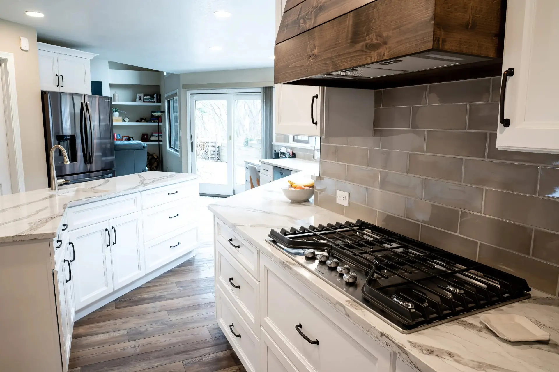 Modern kitchen with white cabinets, marble countertops, and gray subway tiles. A gas stove is on the counter, with a dark wood range hood above. Bright and inviting space.