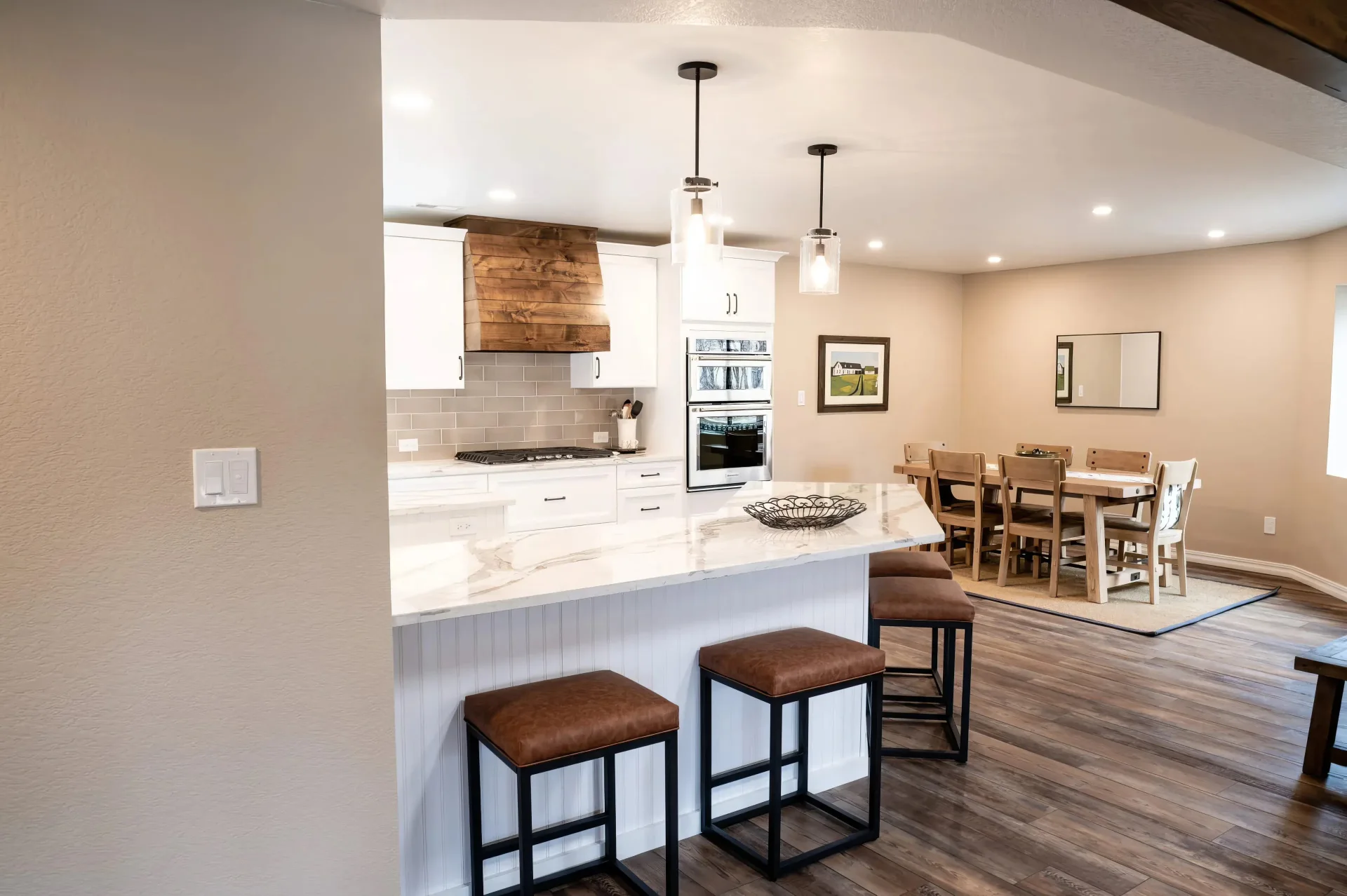 Modern kitchen and dining area with white cabinets, marble island, and brown leather stools. Warm lighting, wooden accents, and cozy dining table.