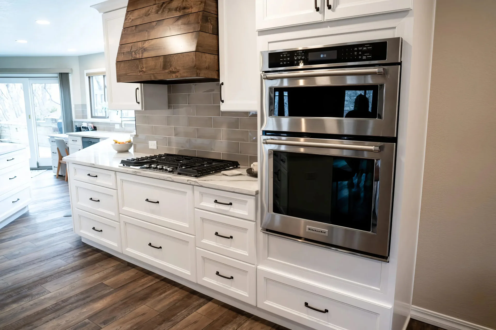 Modern kitchen with white cabinets, stainless steel appliances, and wood accents. A double oven and stovetop sit under a rustic wooden hood. Bright, inviting space.