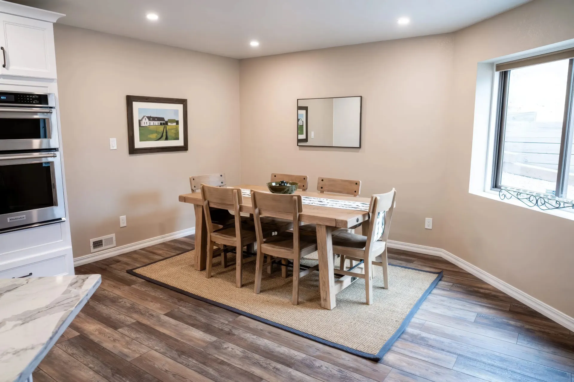 Spacious dining area with a wooden table and six chairs on a rug. Beige walls adorned with a framed art piece and a mirror. Bright and inviting.