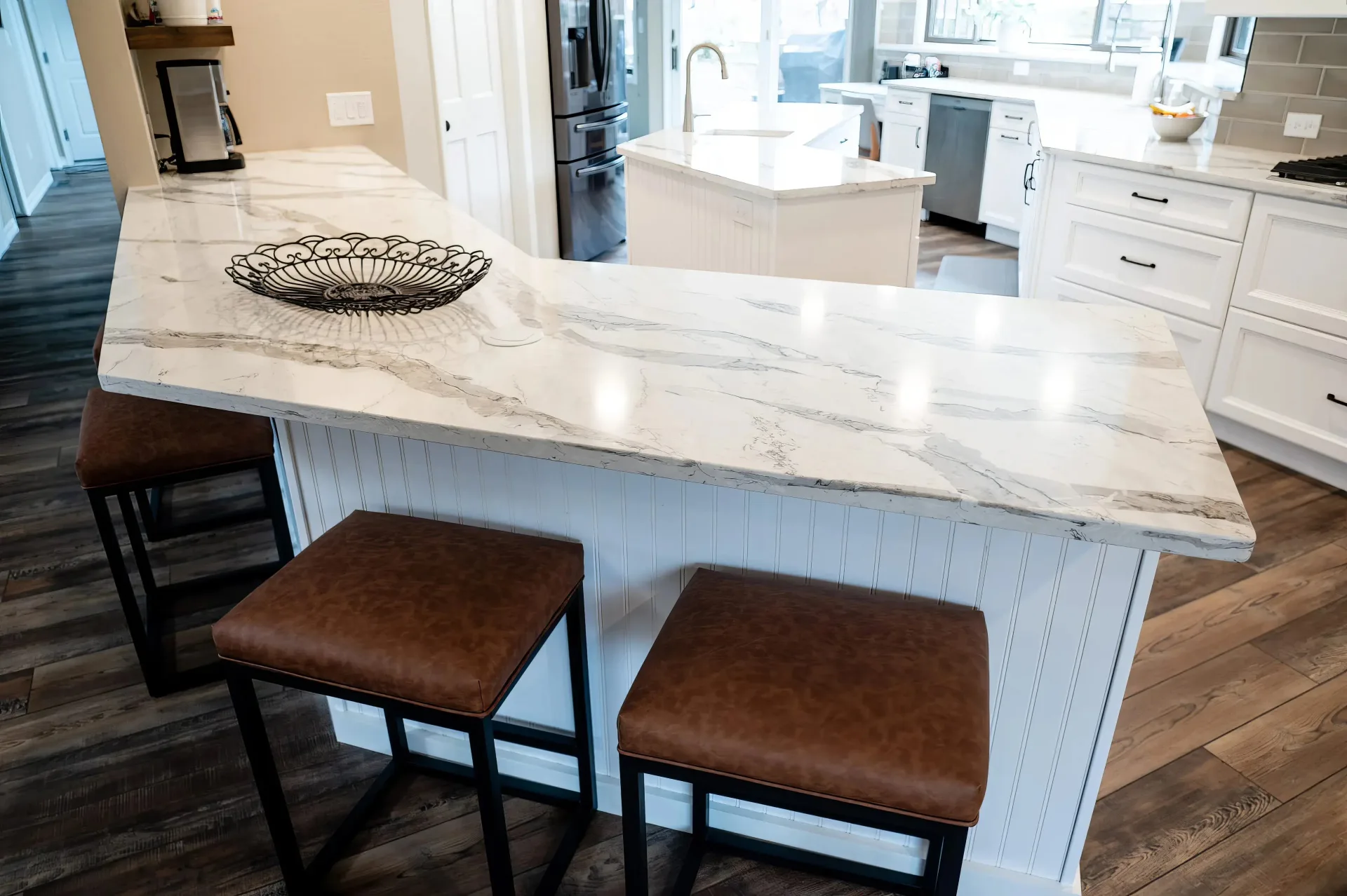 A modern kitchen island with a marble countertop, three brown leather stools, and a decorative metal basket. The space is bright, with wood flooring and white cabinets, conveying a clean and inviting atmosphere.