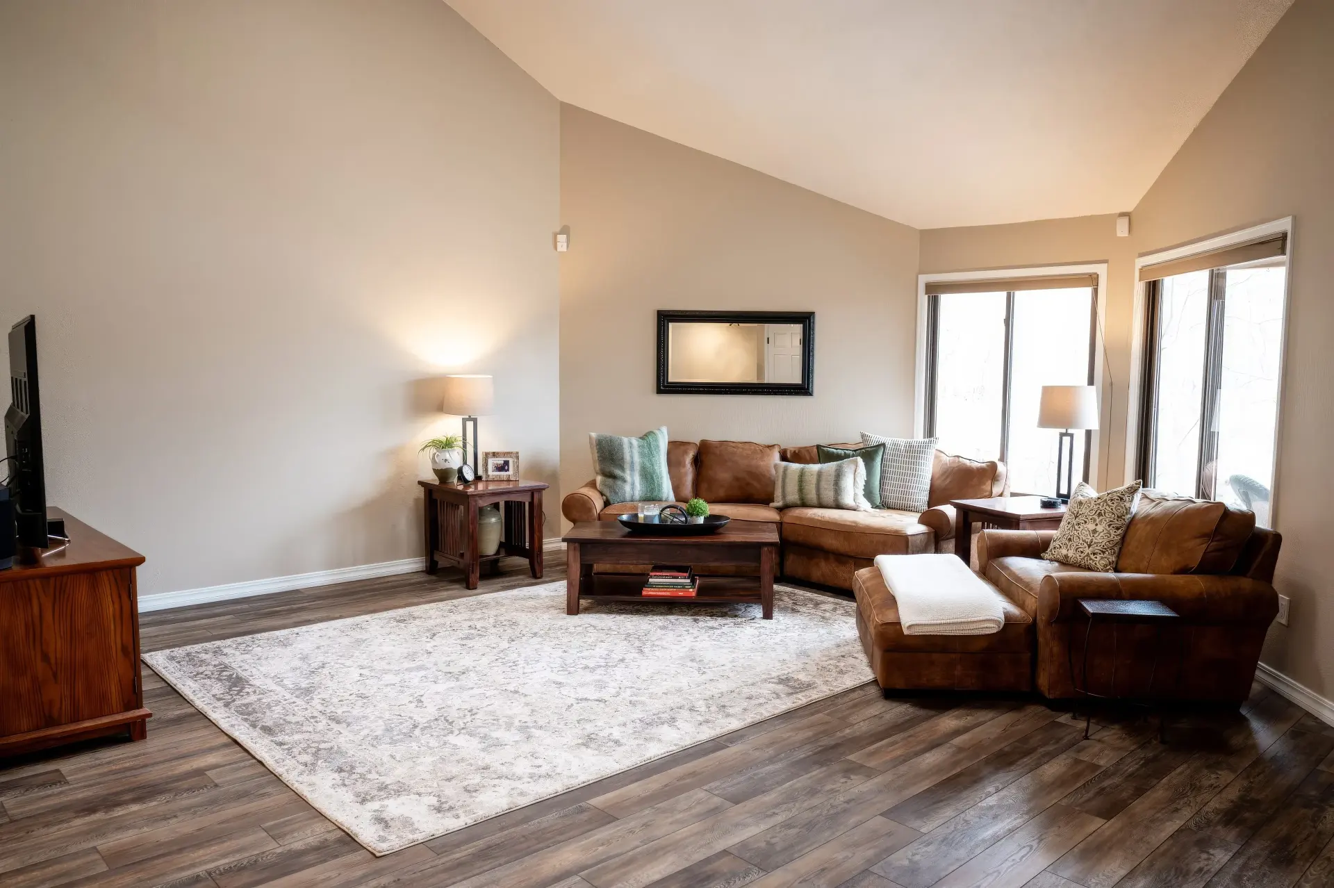 A cozy living room with a high ceiling, featuring a brown leather sectional, armchair, and ottoman on a patterned rug. Soft lighting and large windows create a warm, inviting atmosphere.